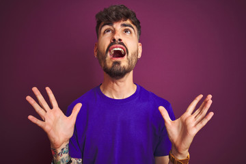Young man with tattoo wearing t-shirt standing over isolated purple background crazy and mad shouting and yelling with aggressive expression and arms raised. Frustration concept.