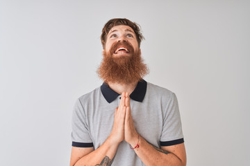 Young redhead irish man wearing grey polo standing over isolated white background begging and...