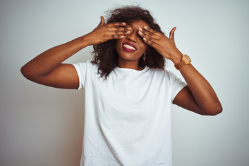 Young african american woman wearing t-shirt standing over isolated white background covering eyes with hands smiling cheerful and funny. Blind concept.