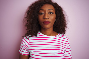 Young african american woman wearing striped t-shirt standing over isolated pink background with serious expression on face. Simple and natural looking at the camera.
