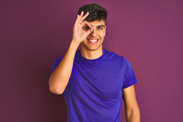 Young indian man wearing t-shirt standing over isolated purple background doing ok gesture with hand smiling, eye looking through fingers with happy face.