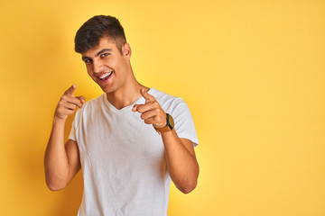 Young indian man wearing white t-shirt standing over isolated yellow background pointing fingers to camera with happy and funny face. Good energy and vibes.