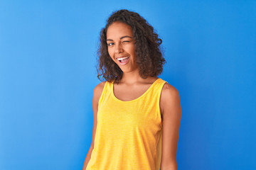 Young brazilian woman wearing yellow t-shirt standing over isolated blue background winking looking...