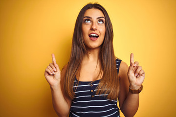 Young beautiful woman wearing striped t-shirt standing over isolated yellow background amazed and surprised looking up and pointing with fingers and raised arms.
