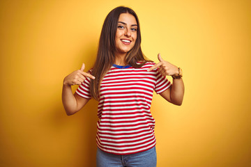 Young beautiful woman wearing striped t-shirt standing over isolated yellow background looking confident with smile on face, pointing oneself with fingers proud and happy.