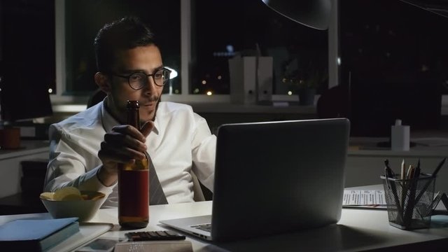 Medium shot of focused Arab businessman in glasses sitting at his desk in dark office and drinking beer from bottle while finishing work on laptop