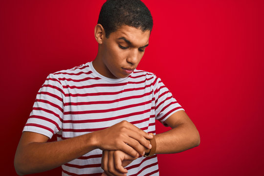 Young handsome arab man wearing striped t-shirt over isolated red background Checking the time on wrist watch, relaxed and confident