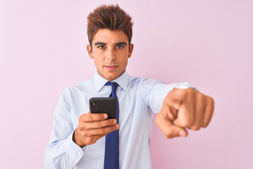 Young handsome businessman using smartphone standing over isolated pink background pointing with finger to the camera and to you, hand sign, positive and confident gesture from the front