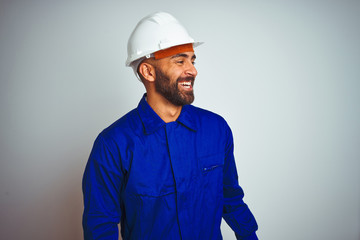 Handsome indian worker man wearing uniform and helmet over isolated white background looking away to side with smile on face, natural expression. Laughing confident.