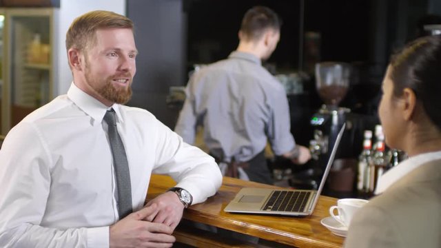 Young caucasian businessman standing at bar counter in cafe and speaking with female colleague while bartender brewing coffee in the background