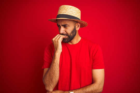 Young handsome indian man wearing t-shirt and hat over isolated red background looking stressed and nervous with hands on mouth biting nails. Anxiety problem.