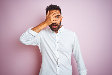Young indian businessman wearing elegant shirt standing over isolated pink background peeking in shock covering face and eyes with hand, looking through fingers with embarrassed expression.