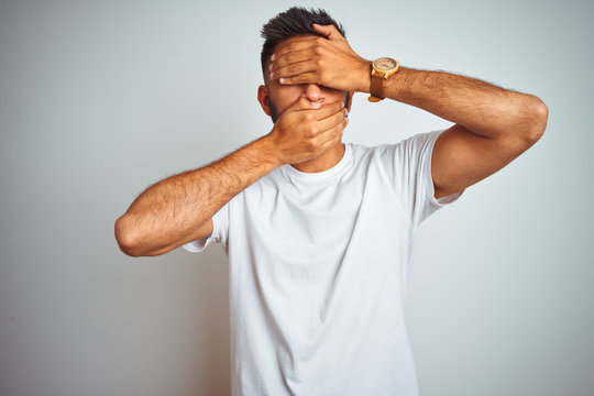 Young indian man wearing t-shirt standing over isolated white background Covering eyes and mouth with hands, surprised and shocked. Hiding emotion
