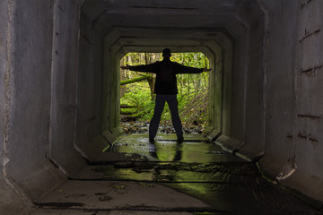 Young man standing on end of concrete stream tunnel in black jacket with backpack and open hand