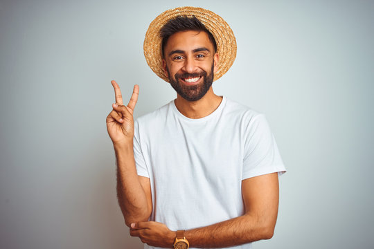 Young indian man on holiday wearing summer hat standing over isolated white background smiling with happy face winking at the camera doing victory sign. Number two.