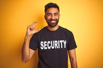 Arab indian hispanic safeguard man wearing security uniform over isolated yellow background smiling and confident gesturing with hand doing small size sign with fingers looking and the camera