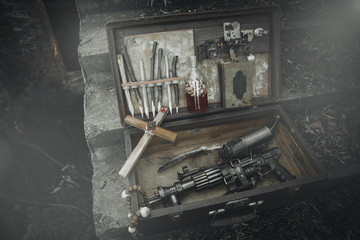 A set of things hunter evil spirits, demons, vampires in an old suitcase. An old book with spells, aspen stakes, tincture of garlic and Holy water, rosary, cross and gun. The view from the side.