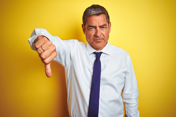 Handsome middle age businessman standing over isolated yellow background looking unhappy and angry showing rejection and negative with thumbs down gesture. Bad expression.