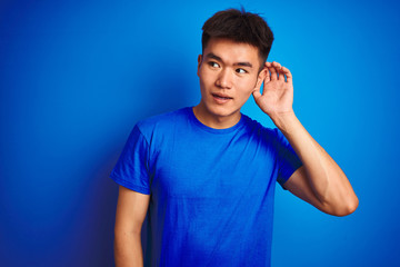 Young asian chinese man wearing t-shirt standing over isolated blue background smiling with hand over ear listening an hearing to rumor or gossip. Deafness concept.