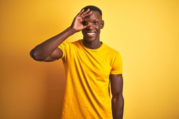 Fototapeta na wymiar Young african american man wearing casual t-shirt standing over isolated yellow background doing ok gesture with hand smiling, eye looking through fingers with happy face.
