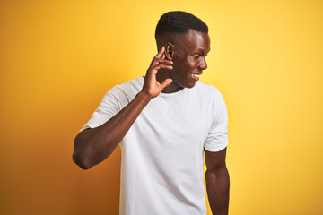 Young african american man wearing white t-shirt standing over isolated yellow background smiling with hand over ear listening an hearing to rumor or gossip. Deafness concept.