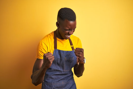 African american bartender man wearing apron standing over isolated yellow background very happy and excited doing winner gesture with arms raised, smiling and screaming for success