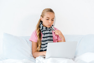 sick kid holding digital thermometer in mouth and looking at laptop