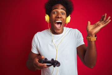 Afro gamer man playing video game using joystick headphones over isolated red background very happy and excited, winner expression celebrating victory screaming with big smile and raised hands