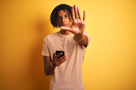 Afro american man with dreadlocks using smartphone over isolated yellow background with open hand doing stop sign with serious and confident expression, defense gesture