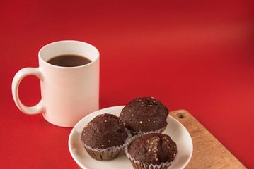 A white Cup of coffee stands on a wooden Board. Chocolate cupcakes are on the plate. Red background.
