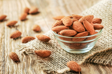 Almonds in glass bowl with green leaves. Almond nuts on wooden table with jute.