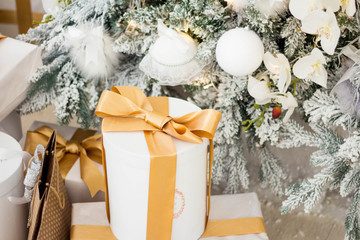 New Year's gold gifts lie under the Christmas tree. Large gift boxes are white with gold ribbon on the floor.