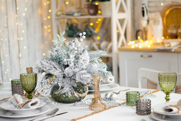 Beautiful served table with decorations, candles with glaze on white tablecloth on the background of the kitchen with people. Living room decorated with lights and Christmas tree.