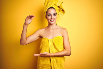 Beautiful woman wearing shower towel on body and head over yellow isolated background gesturing with hands showing big and large size sign, measure symbol. Smiling looking at the camera