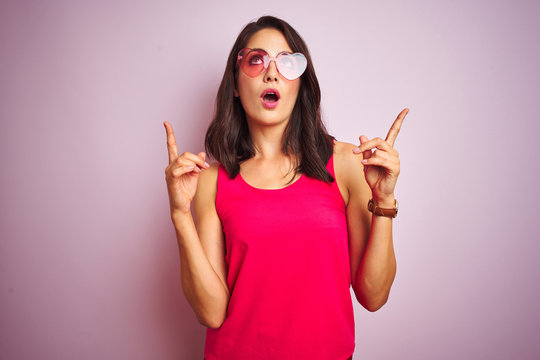 Beautiful woman wearing funny pink heart shaped sunglasses over pink isolated background amazed and surprised looking up and pointing with fingers and raised arms.