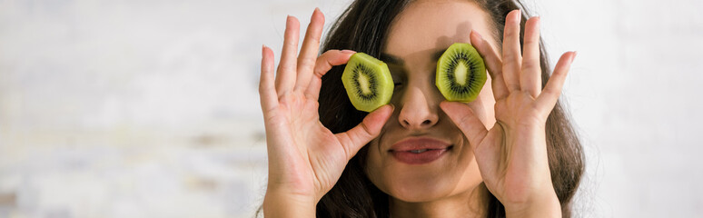 panoramic shot of happy woman holding halves of kiwi fruit while covering eyes