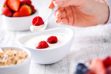 White yogurt with forest fruits and woman hand with yogurt and raspberry on spoon. Red  juicy strawberries in background.