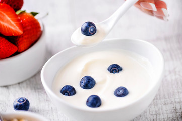 Blueberries in white yogurt. Woman hand holding spoon with one blueberry above white yogurt with forest fruits. Strawberries in background.