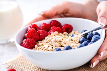 Oat flakeswith forest fruits in white plate on rustic table with jar of milk in backgrund. Woman hands on white plate with oat flakes with raspberries and blueberries.