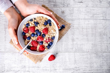 Top view on oat flakes with forest fruits. Raspberries and blueberries in white plate with oat flakes on jute.  Woman hands holding plate of oat flakes with forest fruits.