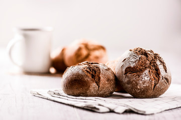 Fresh hot buns and jar of milk in background. Dark rolls of tasty and  beautiful dark bread on white table.
