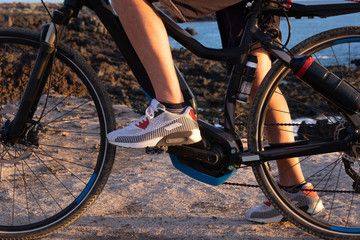 Fototapeta premium Close up of human legs and wheels of an electric bicycle. Footpath along the cliff. Blue water of the sea on background