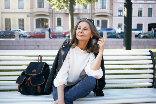 Fashionable elegant middle aged woman relaxing on bench against blurred buildings and cars in background, touching her hair thoughtfully. Rest, leisure, urban lifestyle and relaxation concept