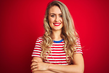 Young beautiful woman wearing stripes t-shirt standing over red isolated background happy face smiling with crossed arms looking at the camera. Positive person.