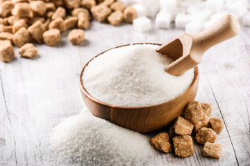 Sugar in bowl with scoop on white rustic table. White sugar and mint leaf on top. Brown and white sugar cubes in back. © Milan
