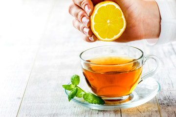 Green tea on white table with green mint leaf. Woman hand squeeze half lemon into tea.