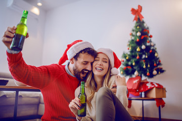 Cheerful caucasian couple with santa hats on heads sitting on the floor, holding beer bottles and celebrating new year. In background is christmas tree. Living room interior.