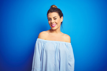 Young beautiful woman wearing bun hairstyle over blue isolated background winking looking at the...