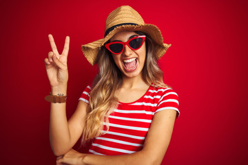 Young beautiful woman wearing sunglasses and summer hat over red isolated background smiling with happy face winking at the camera doing victory sign. Number two.