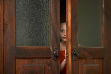A little scared girl is looking through the door slit. A girl is watching someone or something through ajar door. - obrazy, fototapety, plakaty